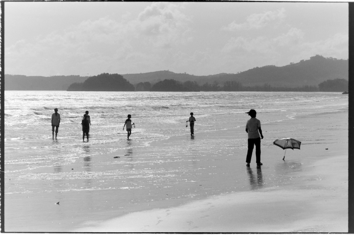 A woman chasing an itinerant umbrella along a beach, her family in the background.